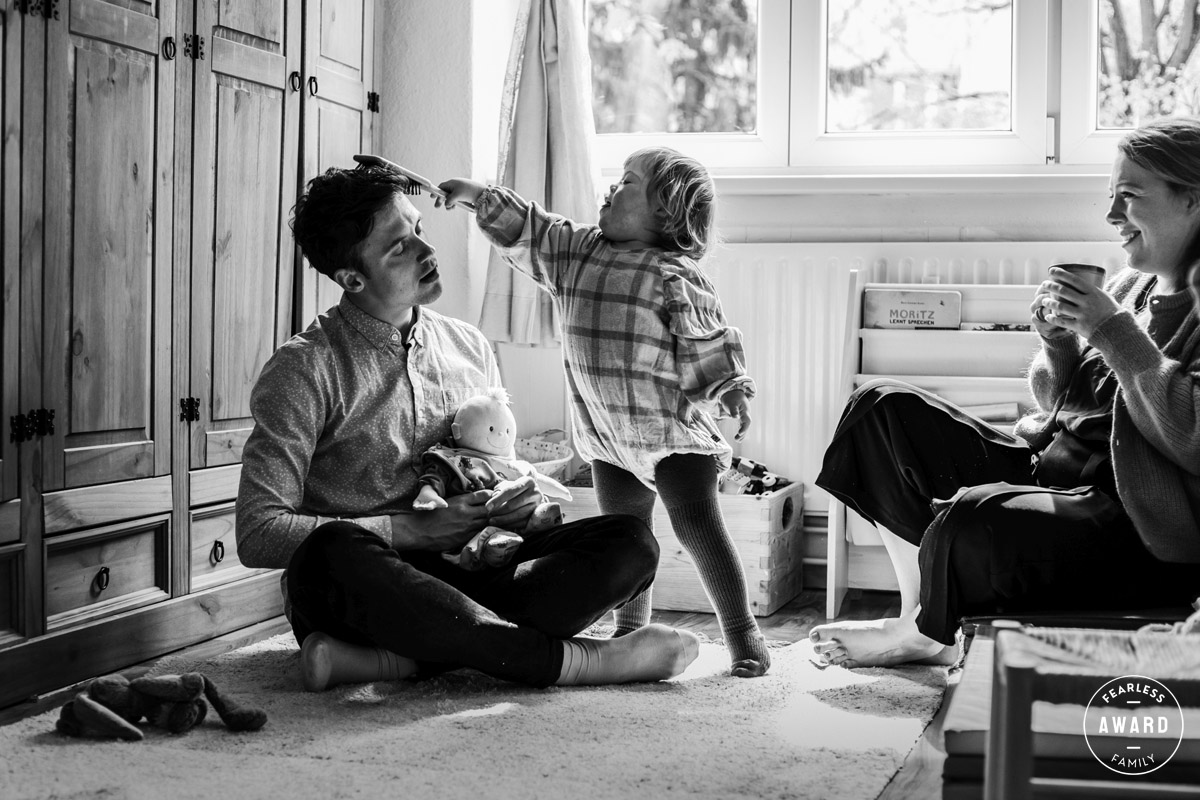 Ein Kind mit Down-Syndrom bürstet seinem Vater die Haare. Der Vater hält eine Babypuppe und hat die Augen geschlossen. Daneben sitzt die Mutter. Sie hält eine Tasse Kaffee und lacht.